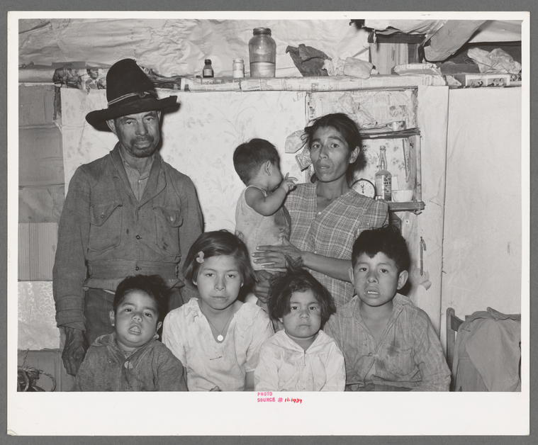 Black and white photo of a Mexican family sitting in their home. Pictured is the mother, father, and five kids.